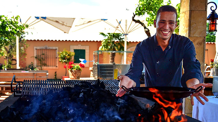young man holds a pan over a grill fire and smiles into the camera